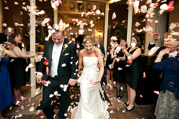 happy bride and groom leaving ceremony as everyone throws light and dark pink rose petals at them - groom is wearing black suit with white vest and tie and bride is wearing white a-line style dress - photo by Houston based wedding photographer Adam Nyholt
